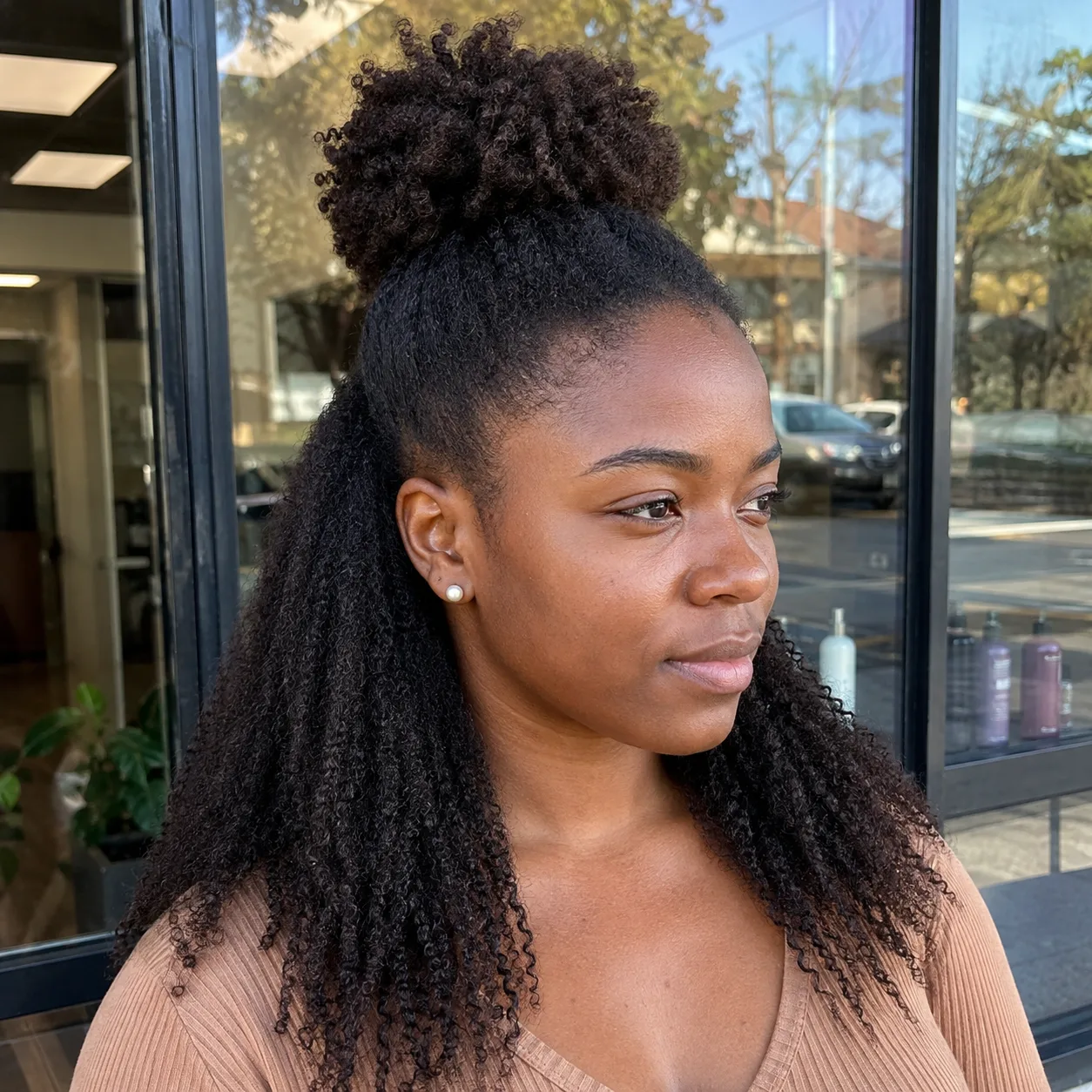 A Black woman outside a salon window with dense dark brown 4C coils, the upper section pulled up into a round puff at the crown and the remaining coils hanging loose past her shoulders