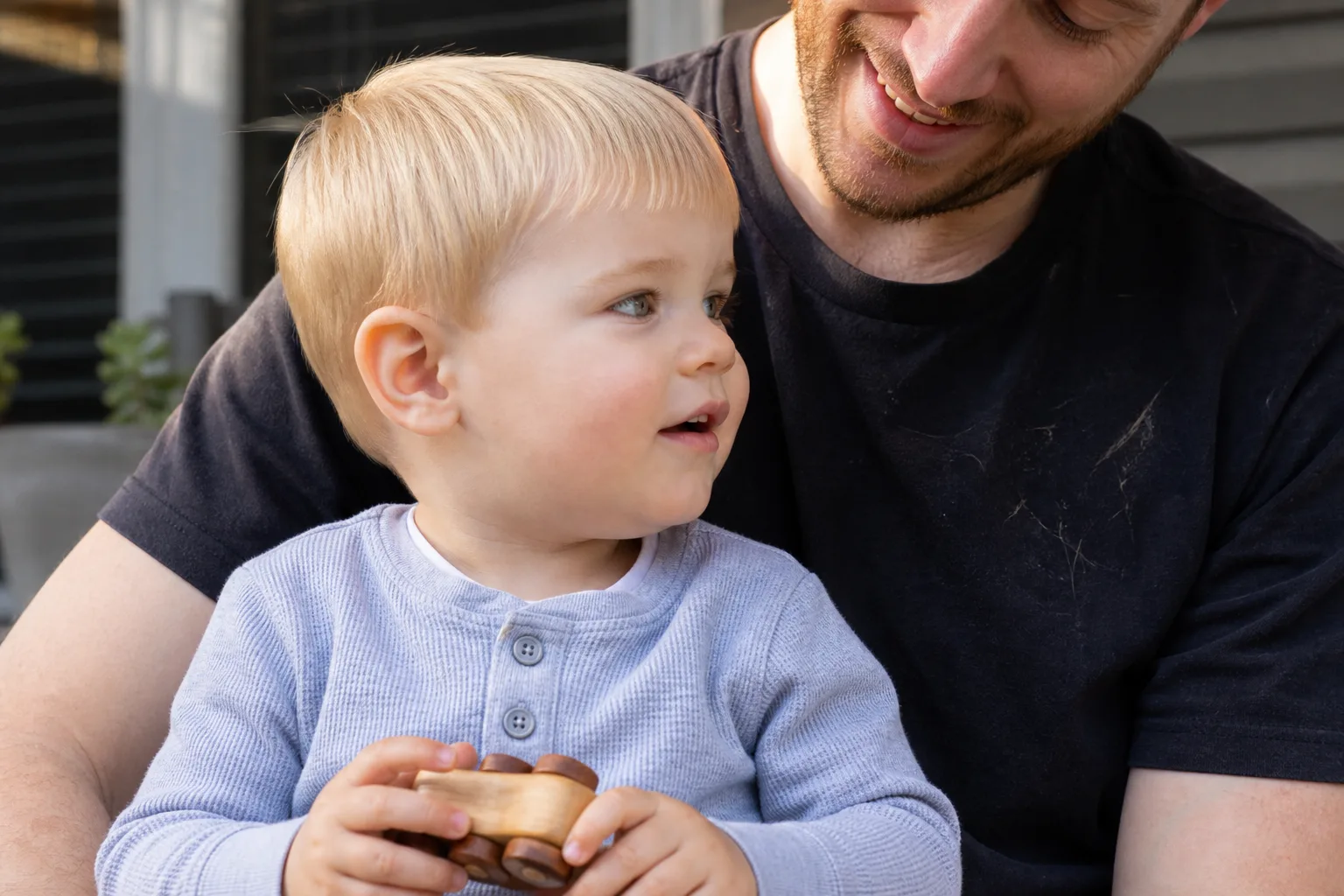 A blonde toddler with a fresh first trim, the hair cut evenly around the ears with a short soft fringe, sitting on a smiling parent's lap on a porch holding a wooden toy car