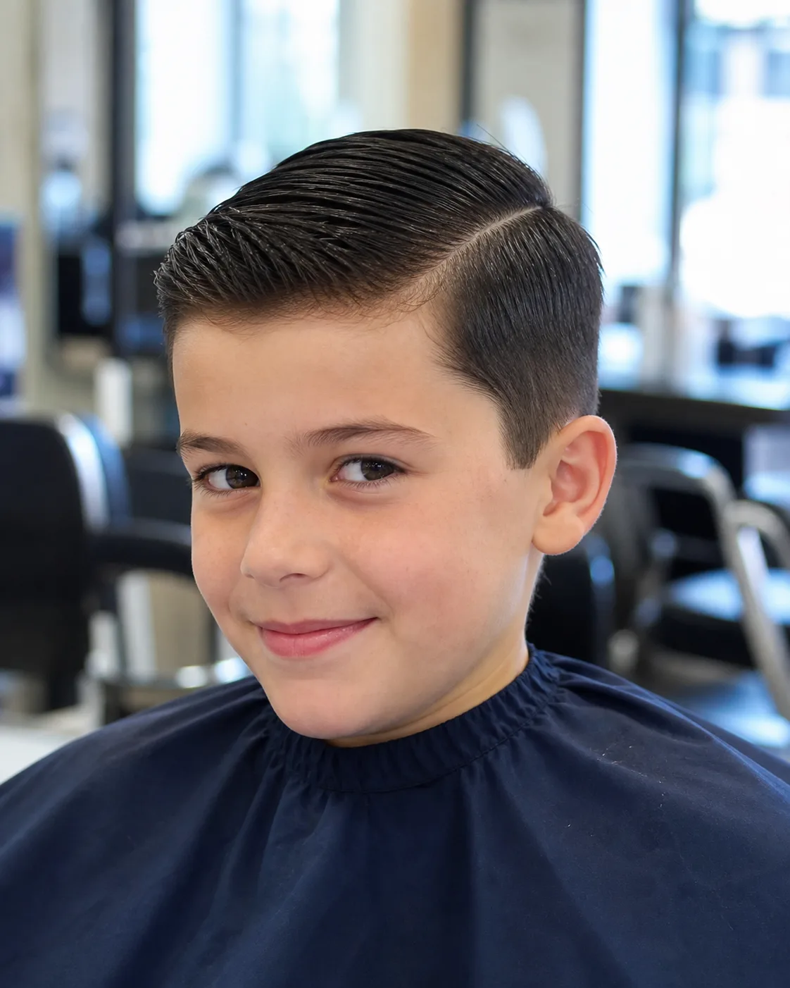 A boy in a navy salon cape with black hair in a short back and sides, the top combed over from a side part