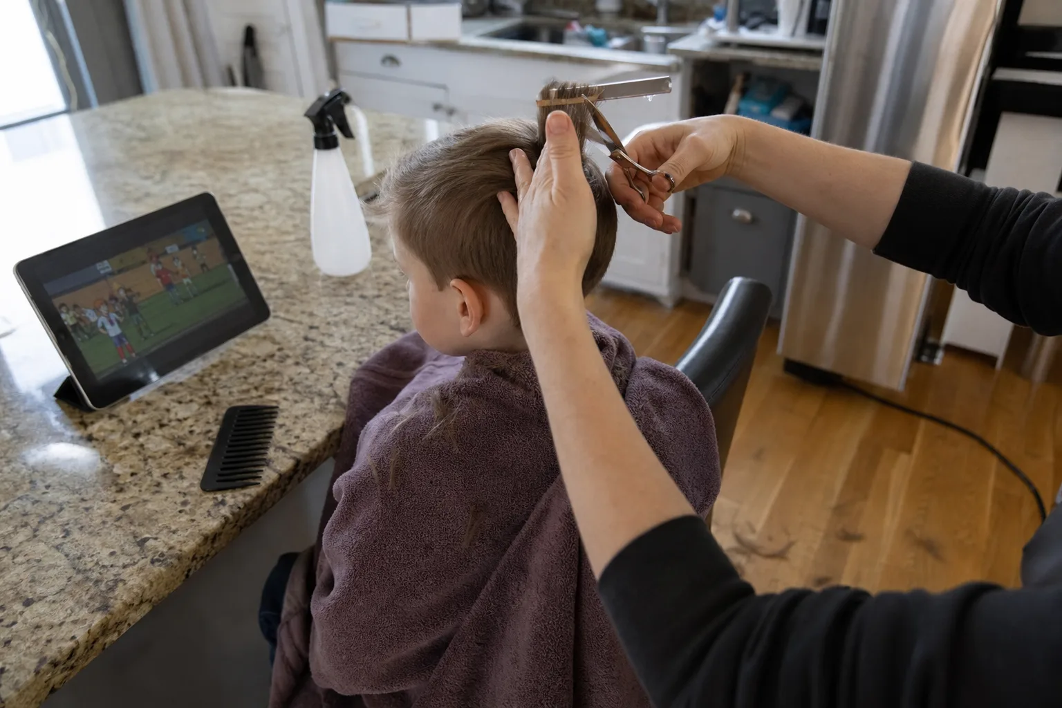 A boy wrapped in a towel on a kitchen stool watches a cartoon on a tablet while an adult holds a section of hair between two fingers and trims the ends with scissors, a spray bottle and comb on the counter