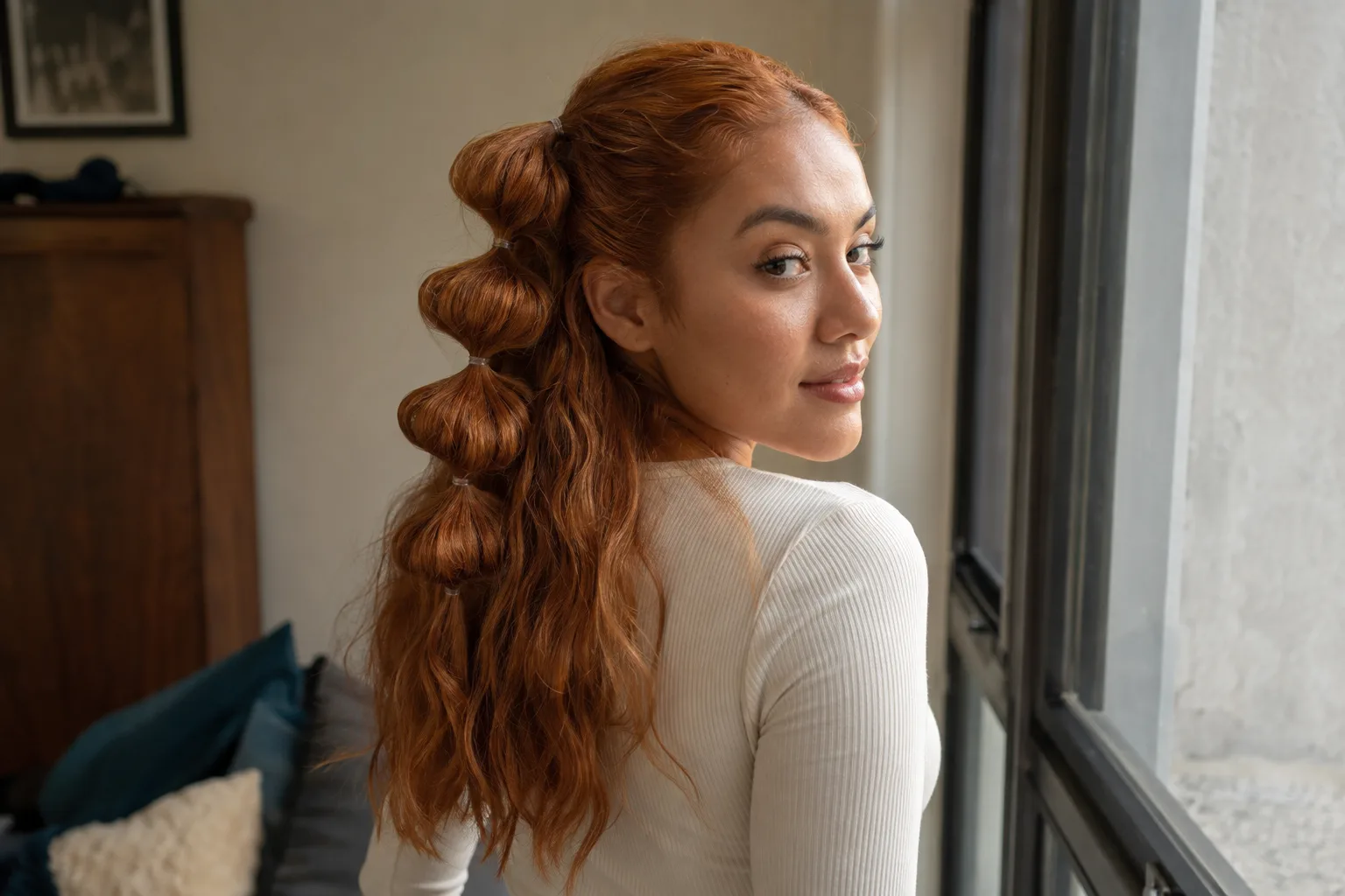 A copper-haired woman by a window, seen from behind, with the top half of her hair gathered into four bubbles and the rest of her waves left loose over her shoulders