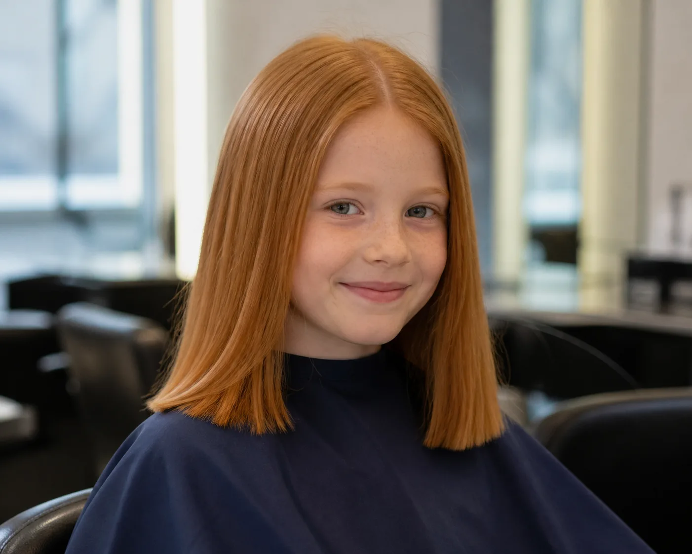 A girl in a navy salon cape with ginger hair in a straight shoulder-length one-length cut with a center part