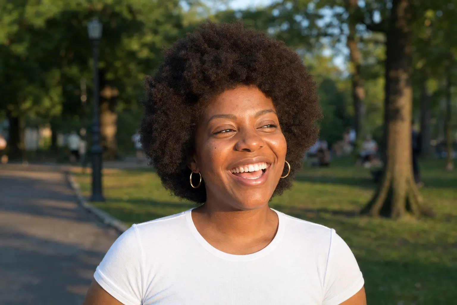 A laughing young woman in a park at golden hour with a full, rounded dark brown afro and gold hoop earrings