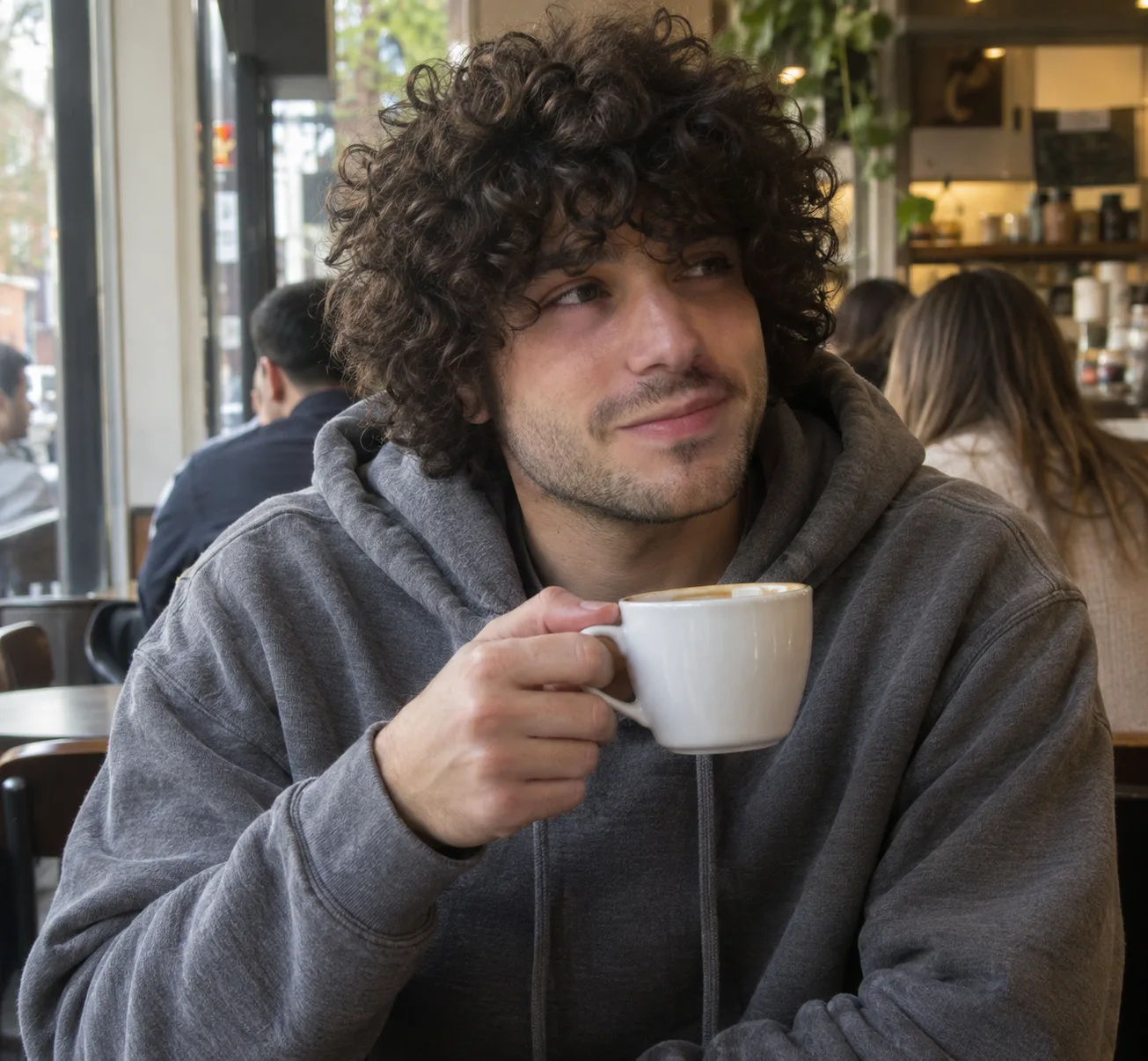 A man with dark brown loose curls covering his ears and falling onto his forehead, holding a coffee cup at a cafe window table