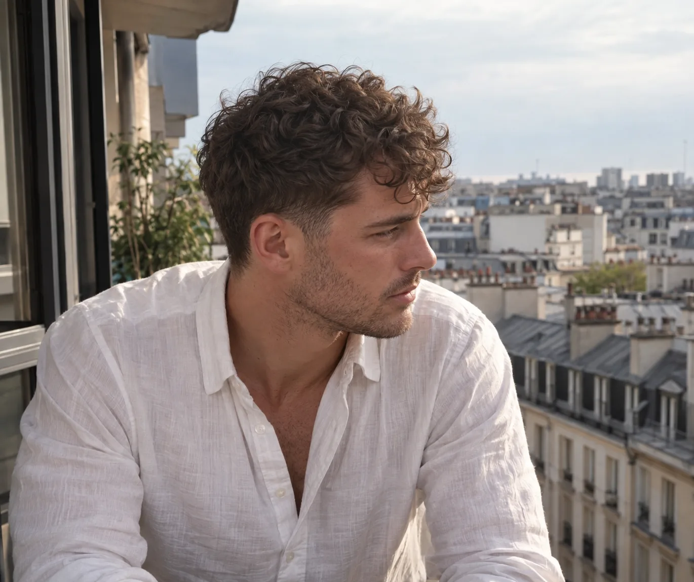A man with short brown curls on top and tapered sides, looking out over city rooftops from a balcony