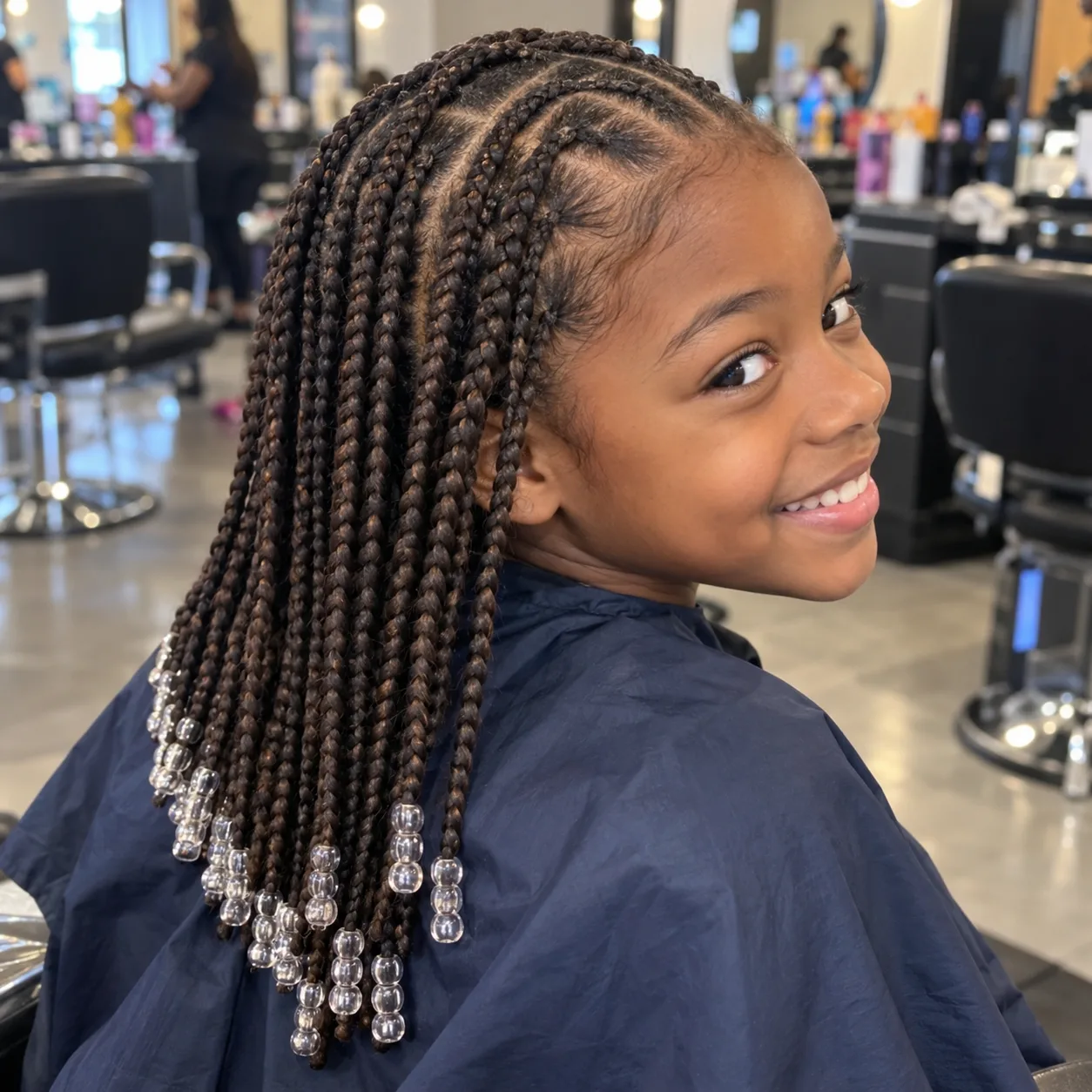 A smiling Black girl in a navy salon cape with dark brown knotless braids past the shoulders, flat at the root, with clear beads on the ends