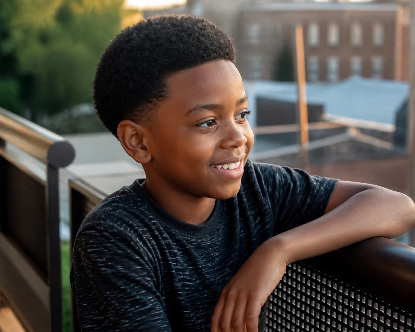 A smiling boy with a rounded black coily top about an inch and a half long and short tapered sides, leaning on a balcony railing in golden evening light