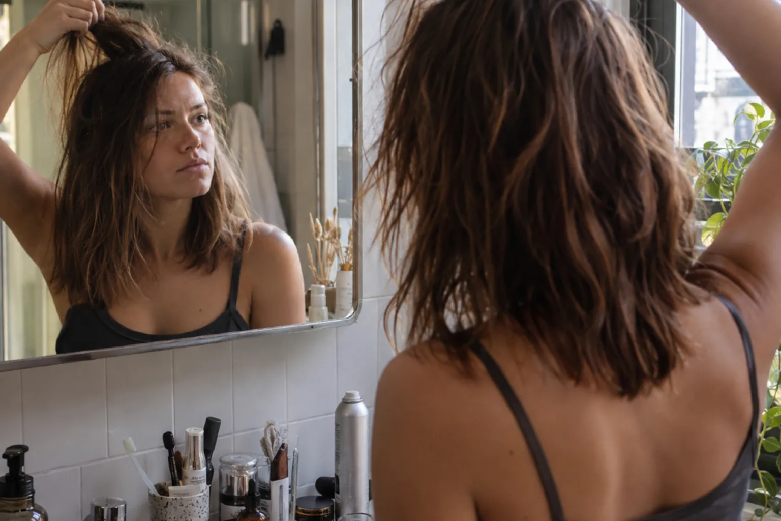 A woman at a bathroom mirror lifting a section of textured second-day hair between two fingers, a can of texture spray on the counter
