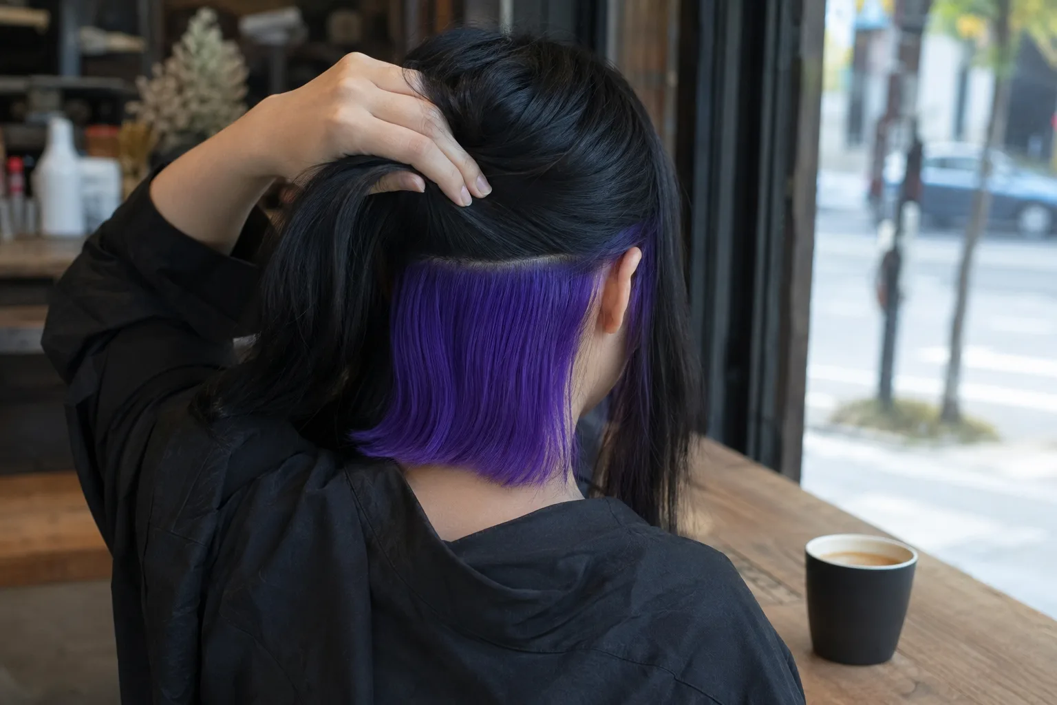 A woman at a cafe window lifts her jet black hair to reveal an electric violet panel underneath, with the outer layer still black
