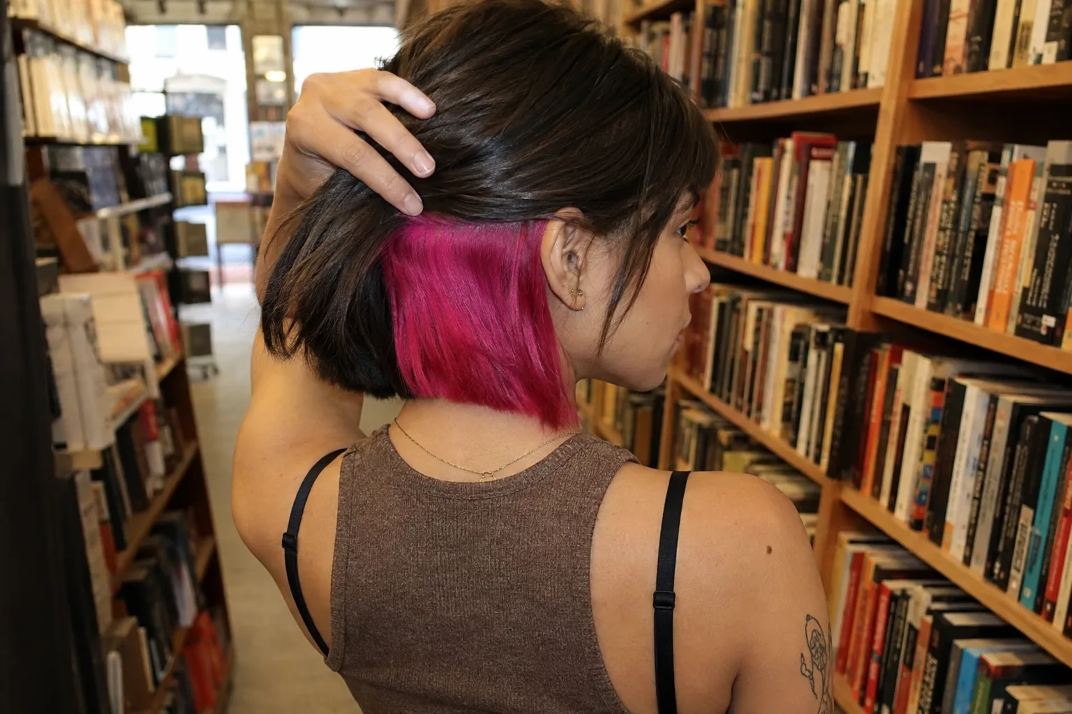 A woman in a bookshop aisle lifts the top layer of her chin-length layered brown cut, showing hot magenta hair underneath