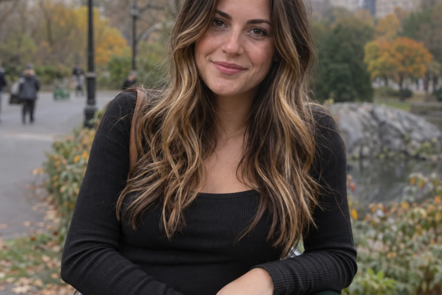 A woman in a city park with very dark brown hair and warm caramel highlights starting below the cheekbone, dark at the roots