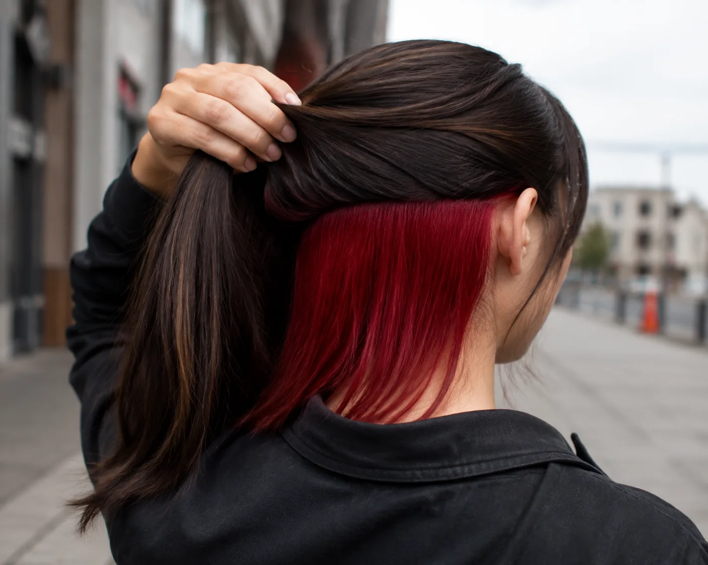 A woman on a city sidewalk lifts her dark brown top layer away from her nape, showing a solid cherry red panel hidden underneath