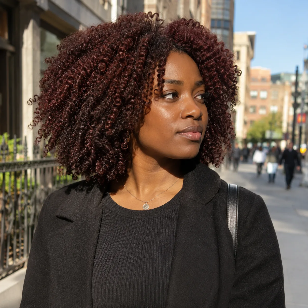 A woman with natural coily burgundy brown hair stands on a sunlit city sidewalk