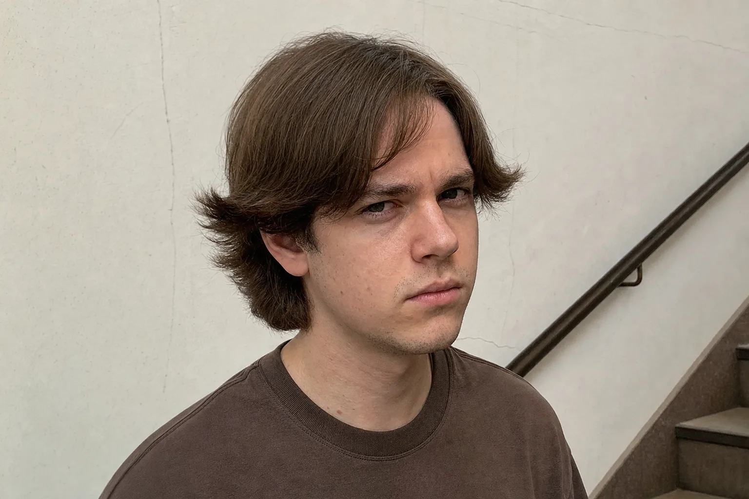 A young man in a stairwell with mid brown hair in the awkward grow-out stage, the side hair kicking out sideways in wings above his ears and a shapeless shelf at the back