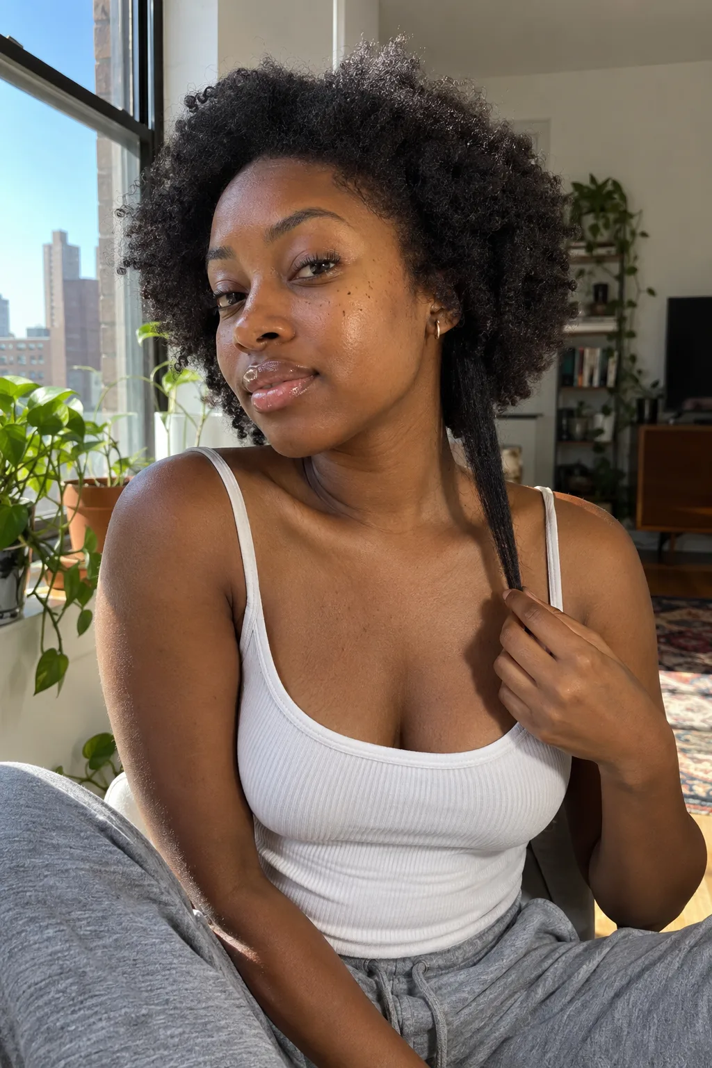A young woman by a sunny apartment window pulling one section of her coily hair straight down past her collarbone while the rest sits as a chin-length afro