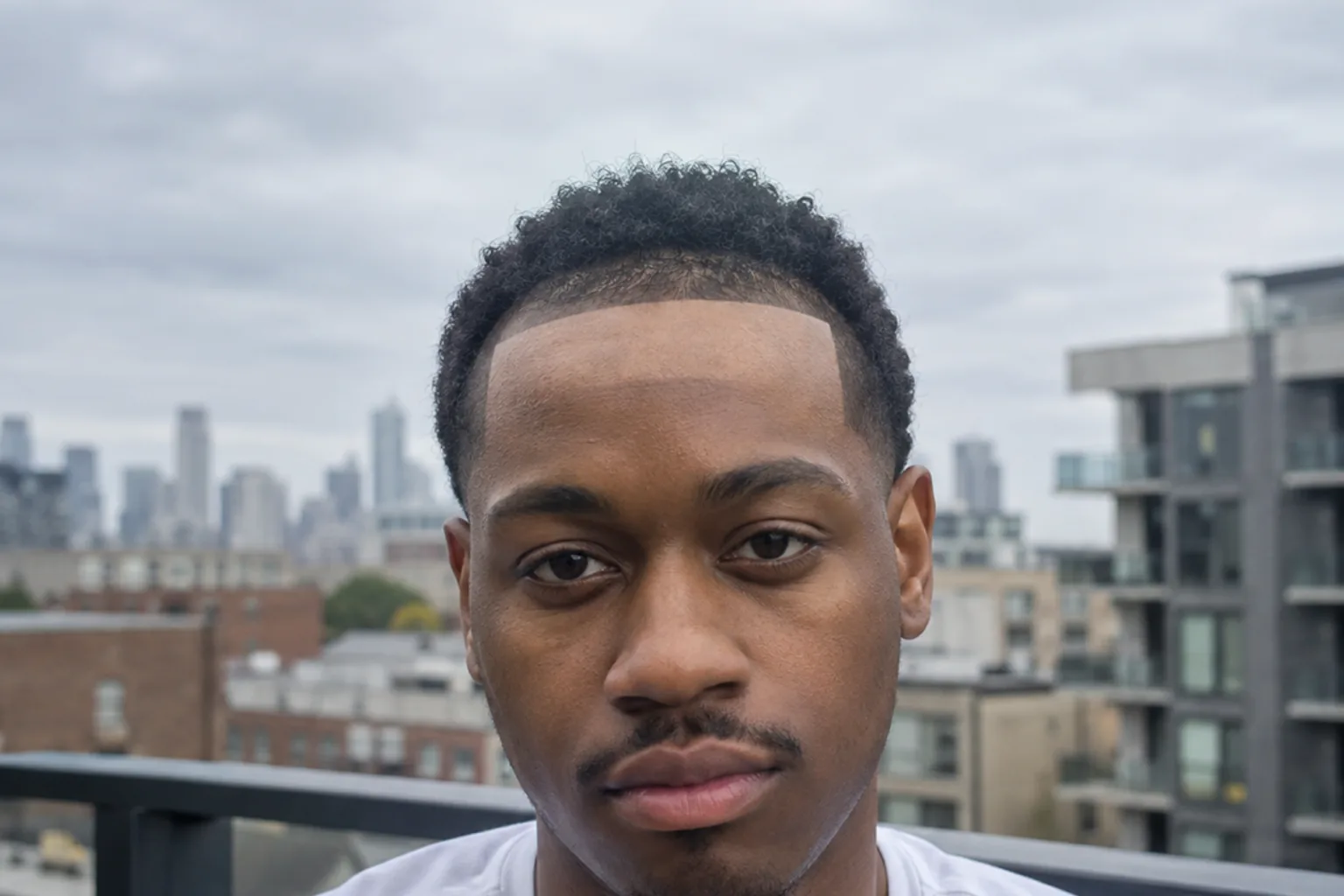 Front view of a young Black man with short black coily hair on an apartment balcony, his front hairline cut into a hard straight edge with sharply squared temple corners