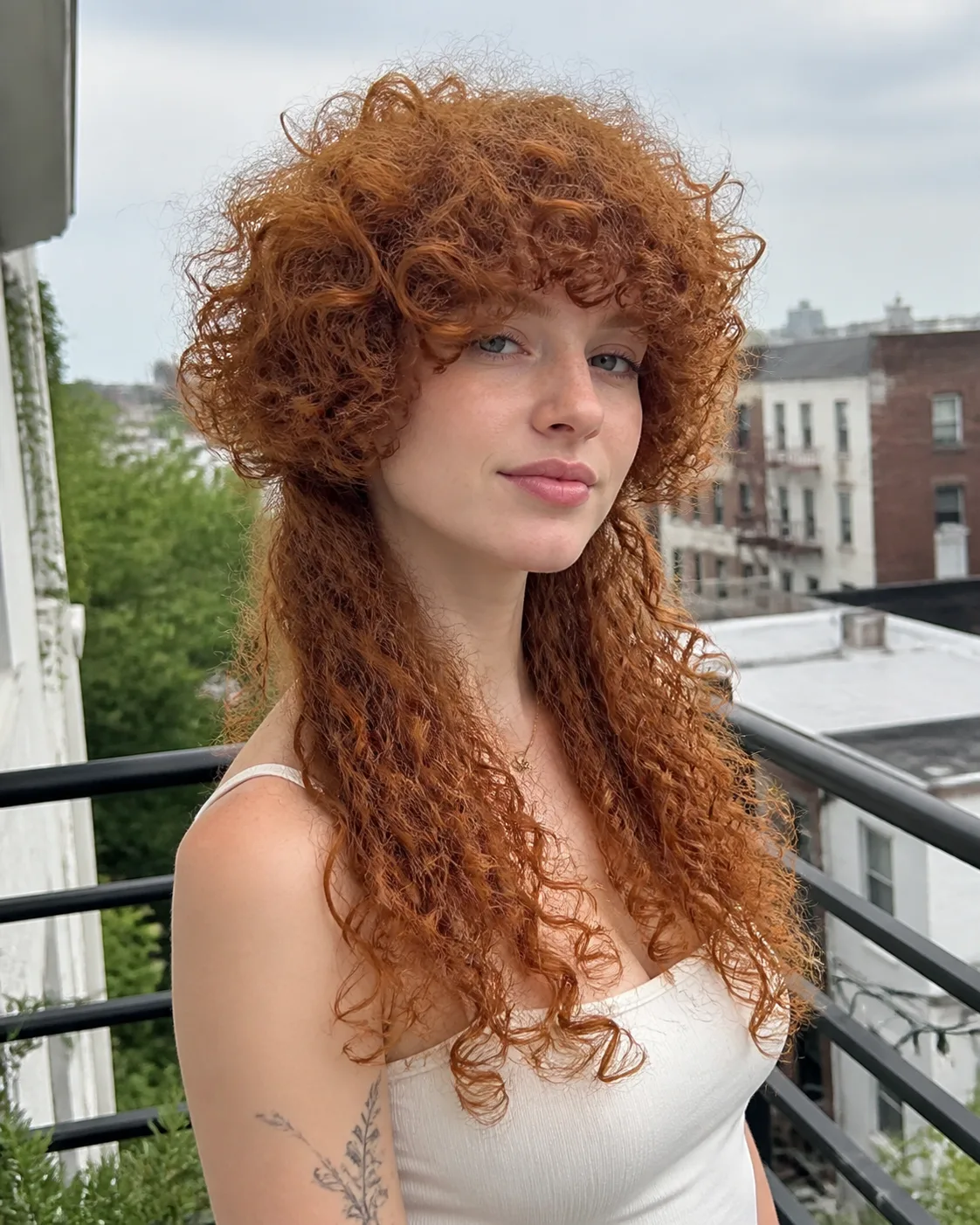 Ginger haired woman on an apartment balcony with a short cap of tight curls sitting above much longer curls down her back