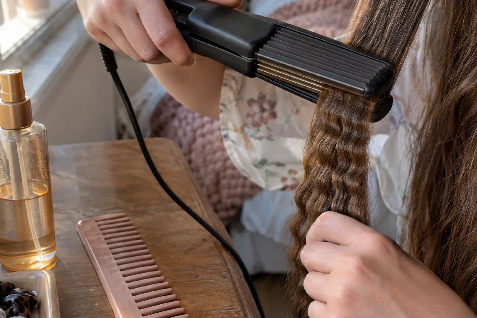 Hands holding a ribbed ceramic crimping iron clamped on a section of mid-brown hair, heat protectant and a wide tooth comb on the table