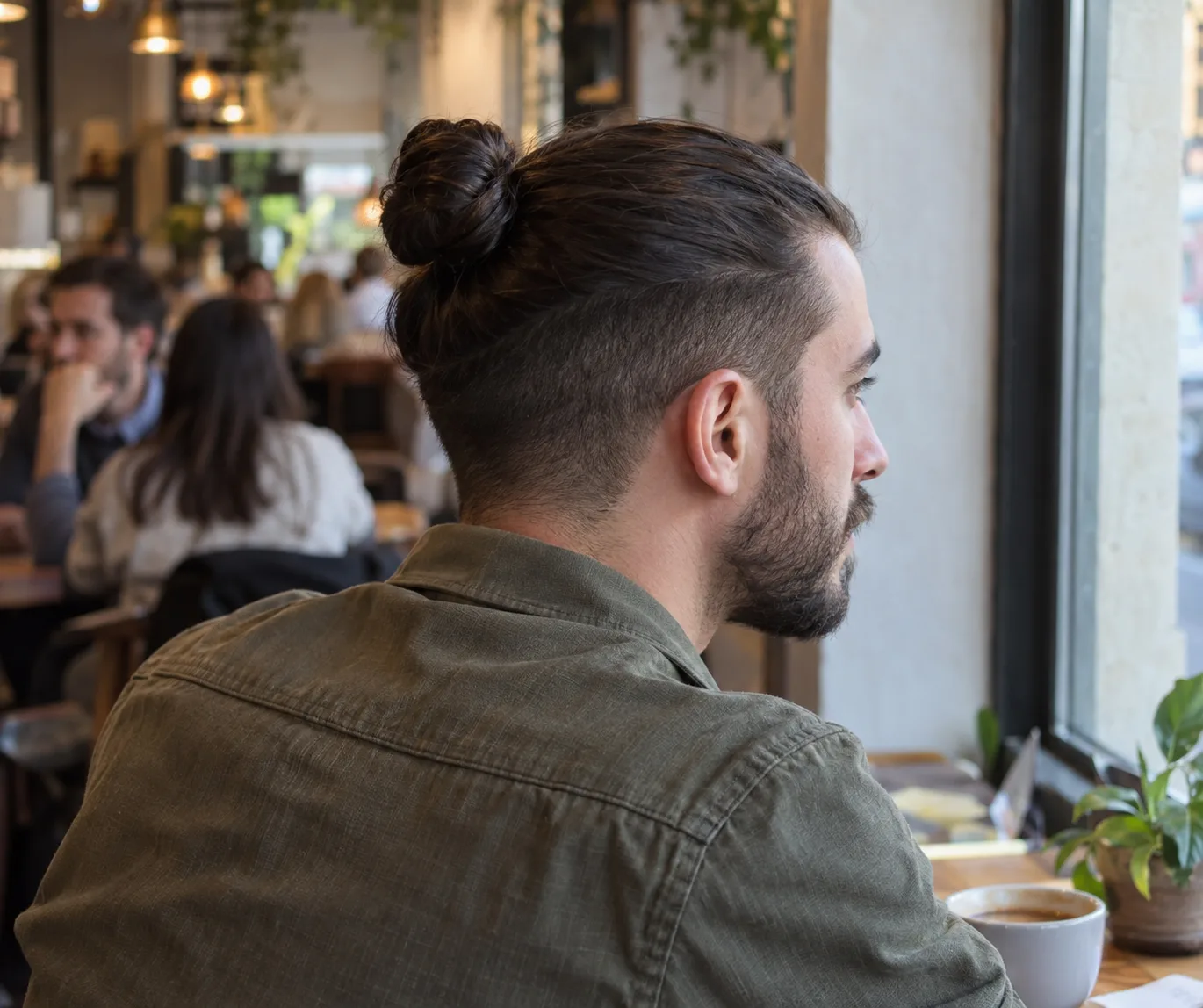 Man at a cafe window with dark hair in a bun on top and short clipped sides and back