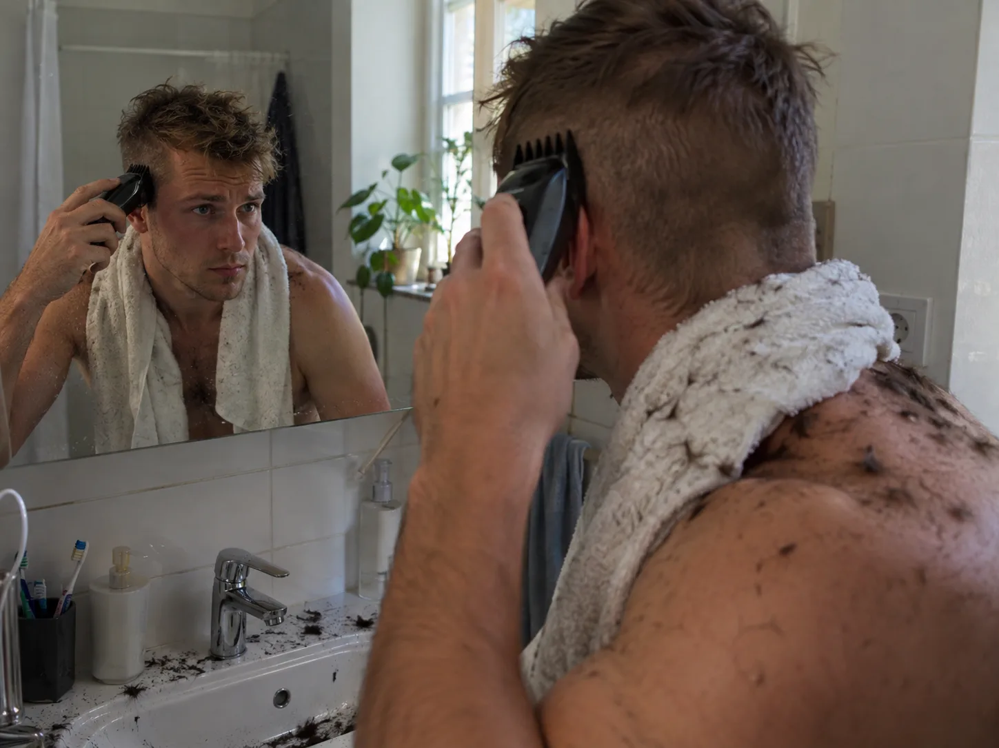 Man cutting his own hair in a bathroom mirror with clippers, sides clipped short and the top still long