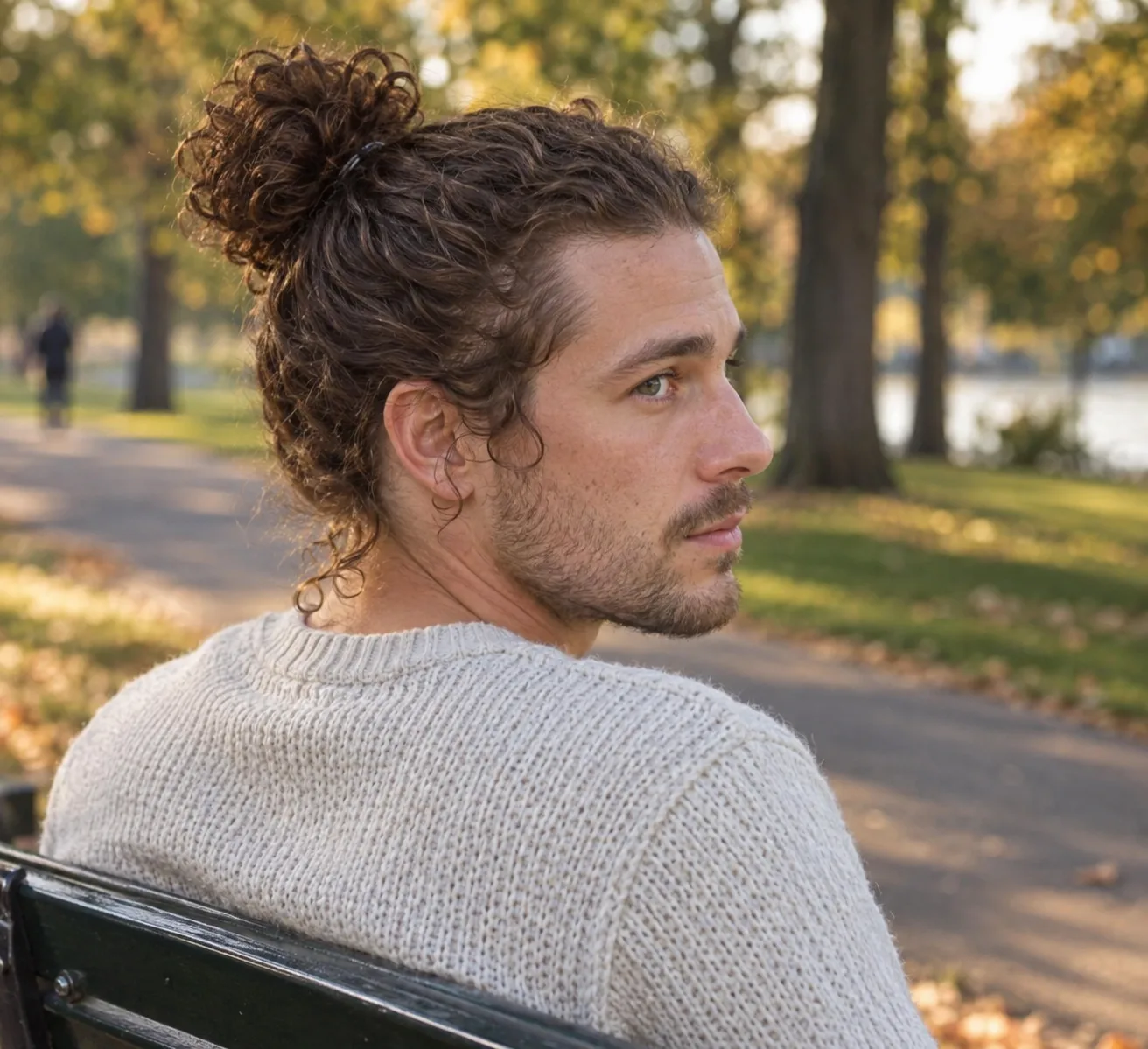 Man in a cream sweater on a park bench with curly brown hair gathered into a loose curly bun