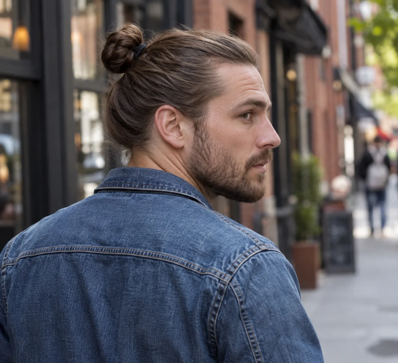 Man in a denim jacket on a city street with his brown hair tied in a bun at the crown