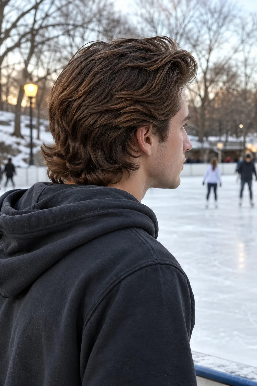 Man in a hoodie seen from behind at an outdoor ice rink in winter, brown hockey flow with thick layers flicking out at the nape