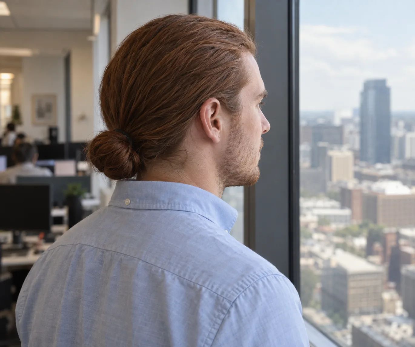 Man in a light blue oxford shirt at an office window wearing an auburn low bun just above his collar