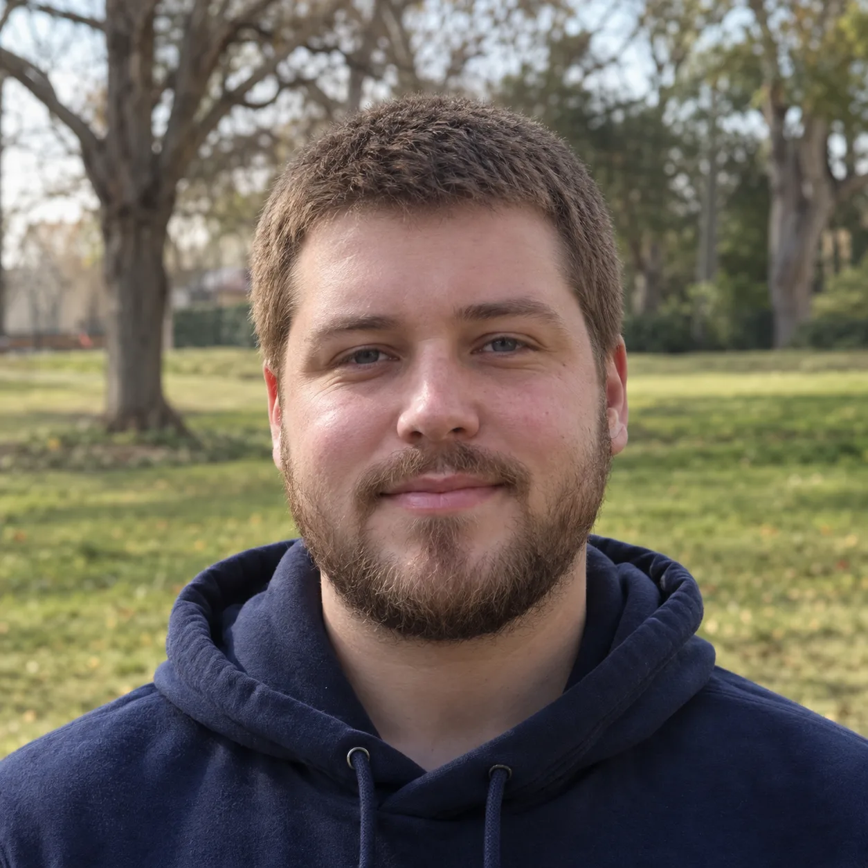 Man with a round face, a short full beard and a light brown 8 guard buzz cut, standing in a park