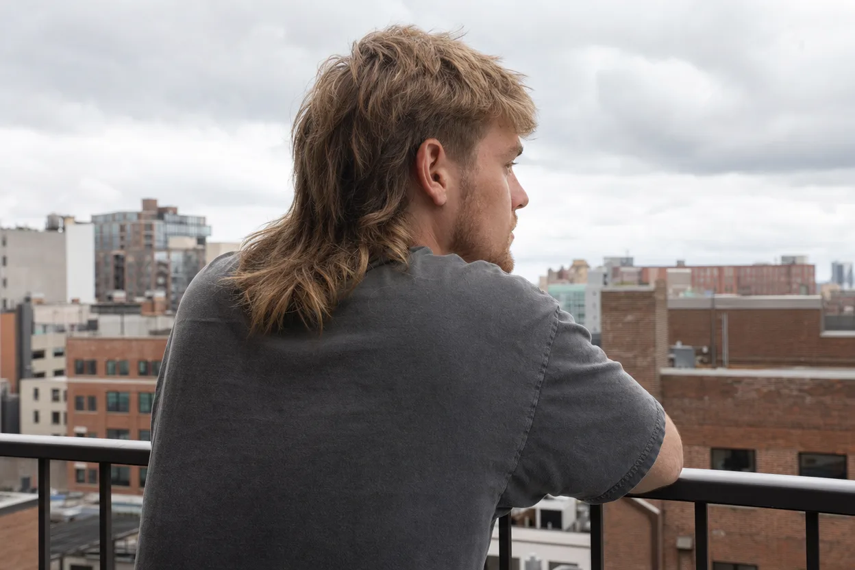 Rear three-quarter view of a young man on a balcony with a blonde modern mullet falling over his collar