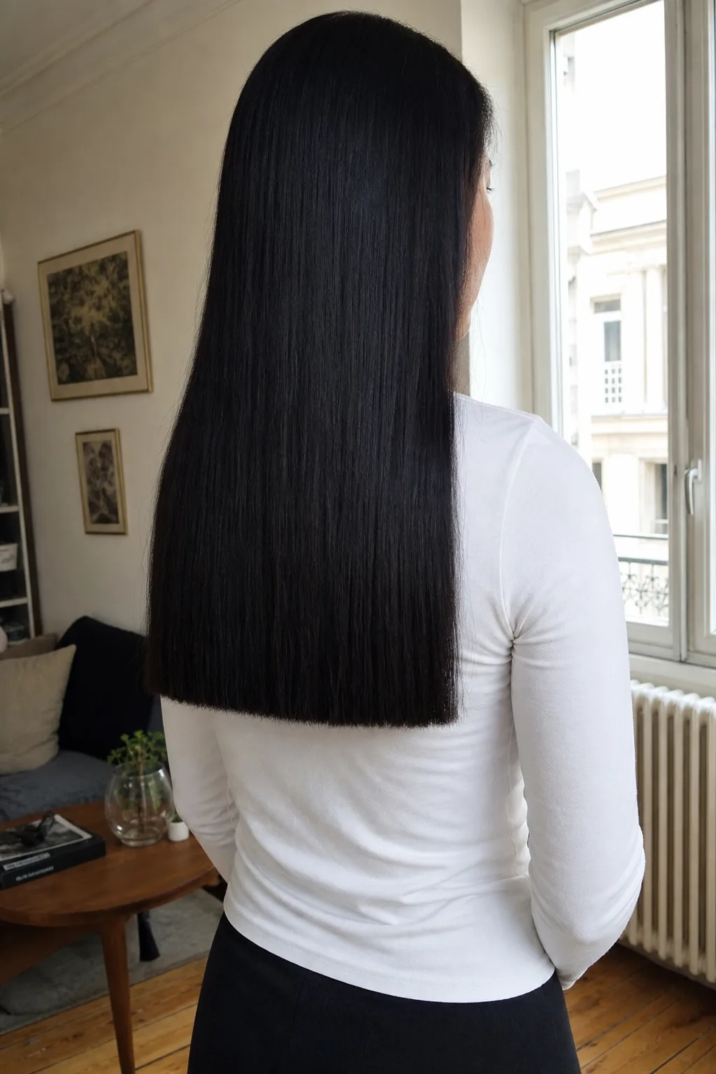 Rear view of a woman by an apartment window, her long glossy black hair hanging flat down her back and ending on one hard straight line across thick blunt ends