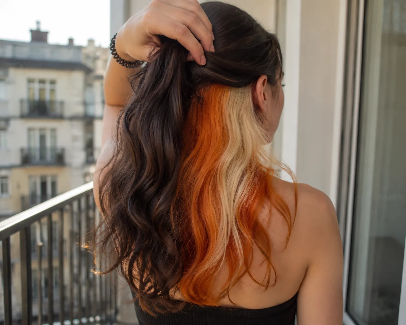 Rear view of a woman on a balcony lifting her dark top layer to reveal orange and blonde panels hidden underneath