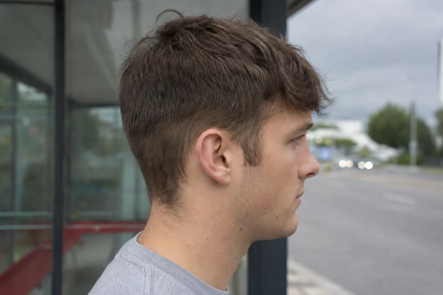 Right side profile of a young man at a bus stop whose fade has grown out, the side filled in to an even soft shadow with no bare skin, a fuzzy nape and no visible line where the short work ended