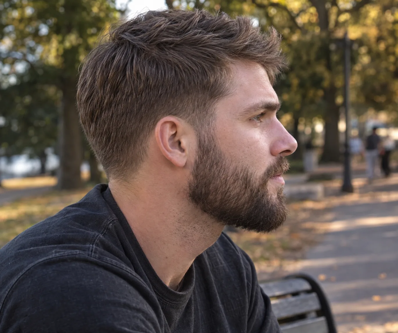 Side profile of a man on a park bench with a low taper fade, the tapered sideburn blending into a short full beard at the same length with no gap