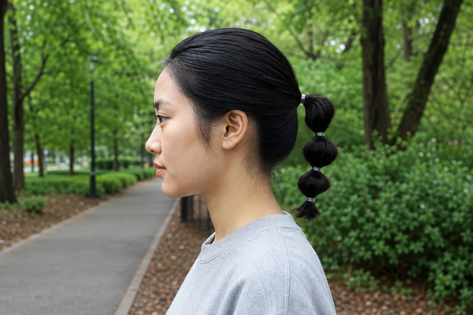 Side profile of a woman in a park with a sleek low bubble ponytail, only four small rounded puffs down a collarbone-length tail