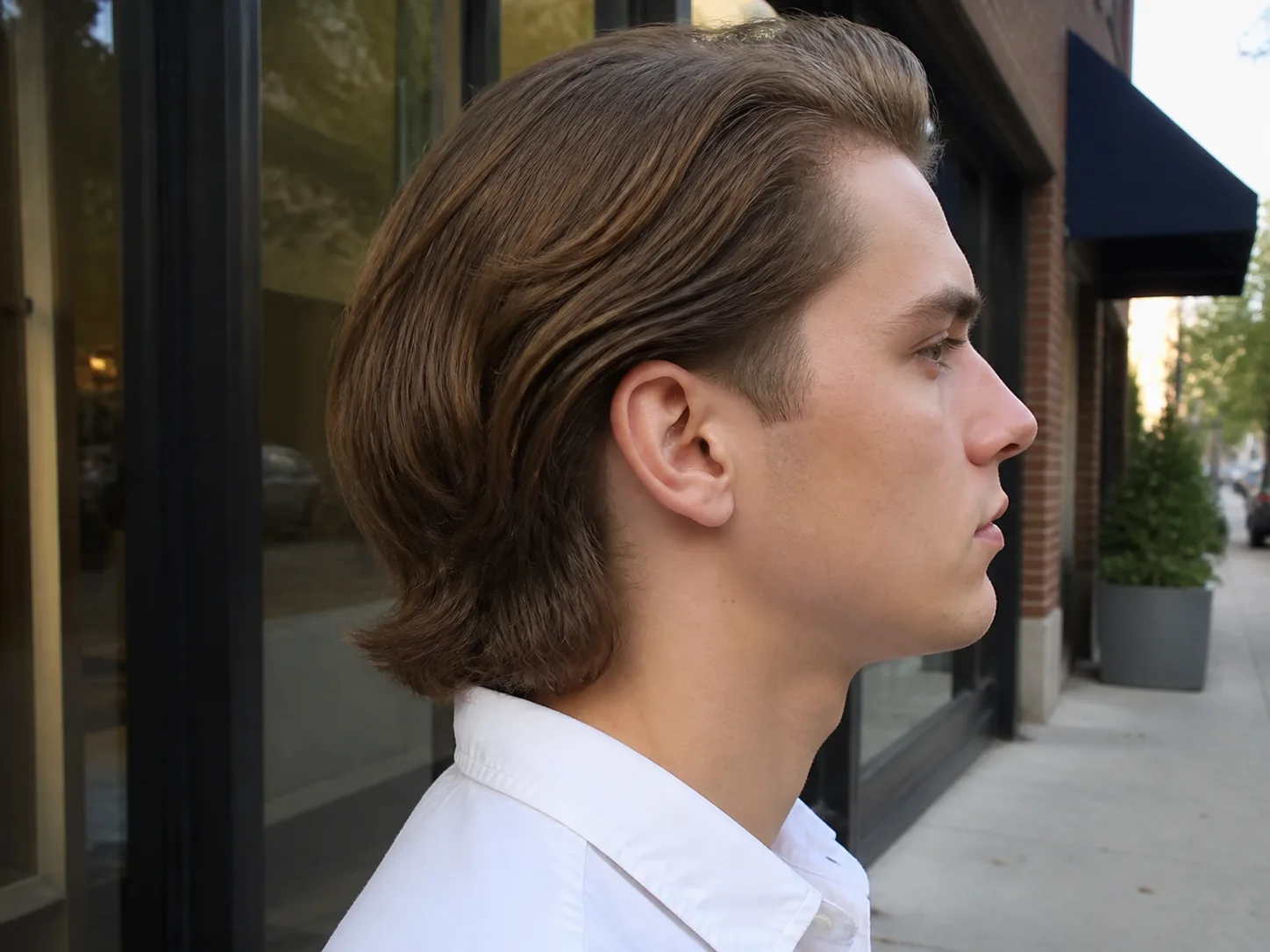 Side profile of a young man with long light brown hair swept back behind his ear and falling to his collar, the low taper visible only as a small tidy graduated area around the ear and sideburn