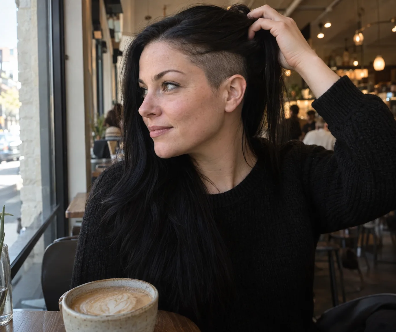 Woman in a cafe lifting her long black hair to show a shaved undercut above her ear