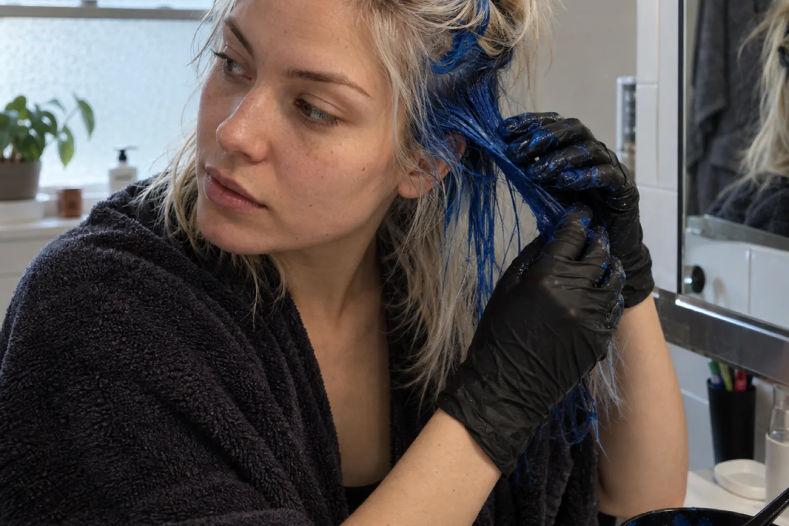 Woman in a dark robe and black gloves working royal blue dye into her blonde hair in a home bathroom