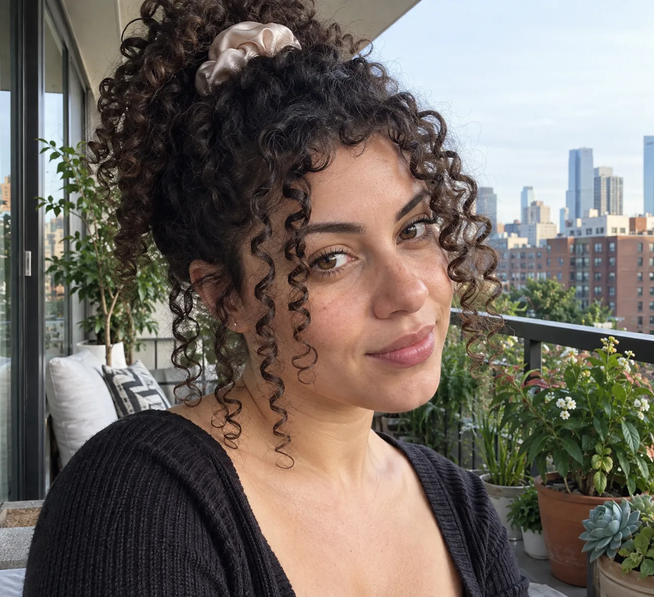 Woman on a city balcony with dark curly hair gathered loosely on top of her head with a satin scrunchie