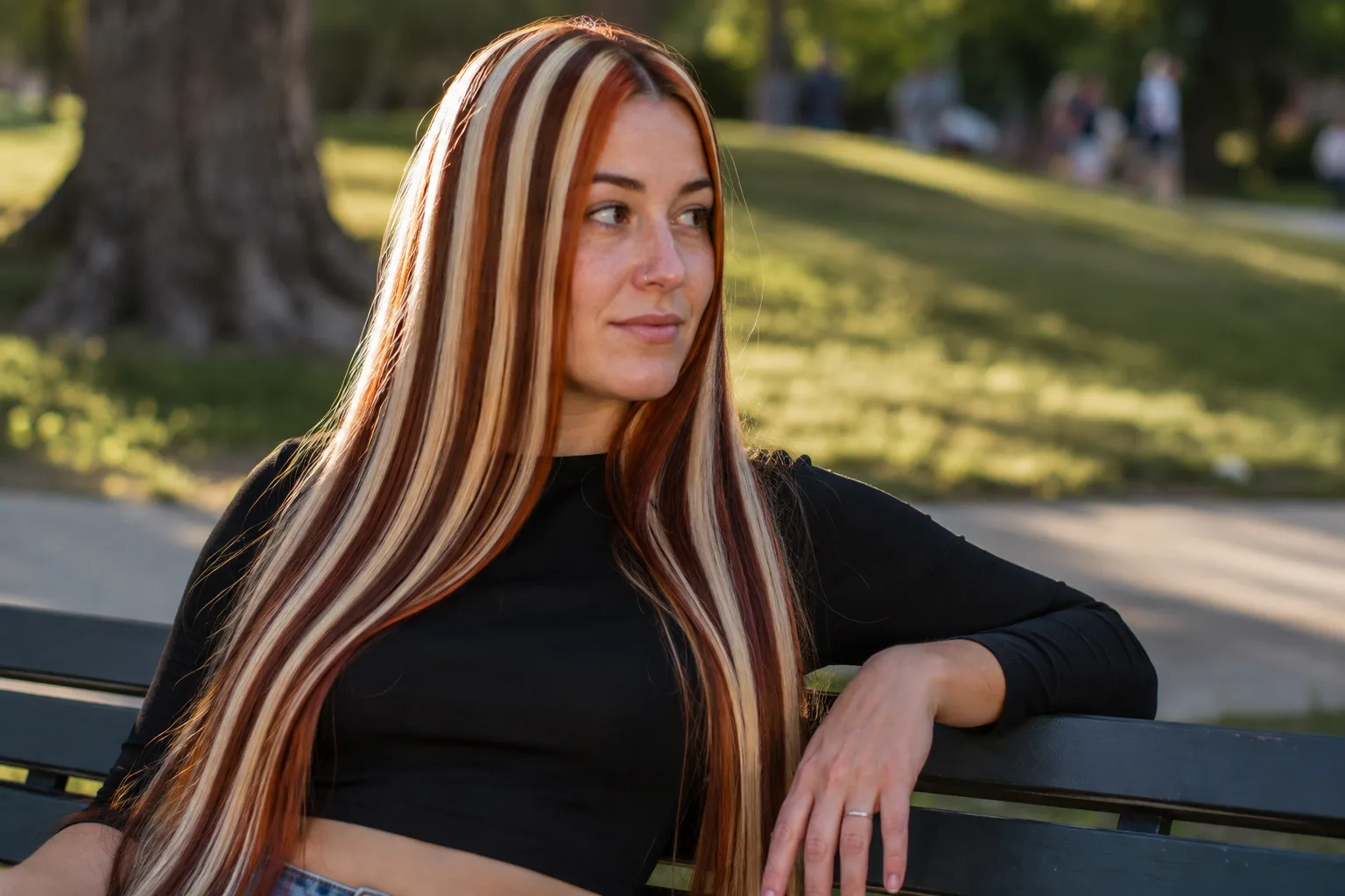 Woman on a park bench with long straight hair dyed in even repeating red and platinum vertical stripes of identical width