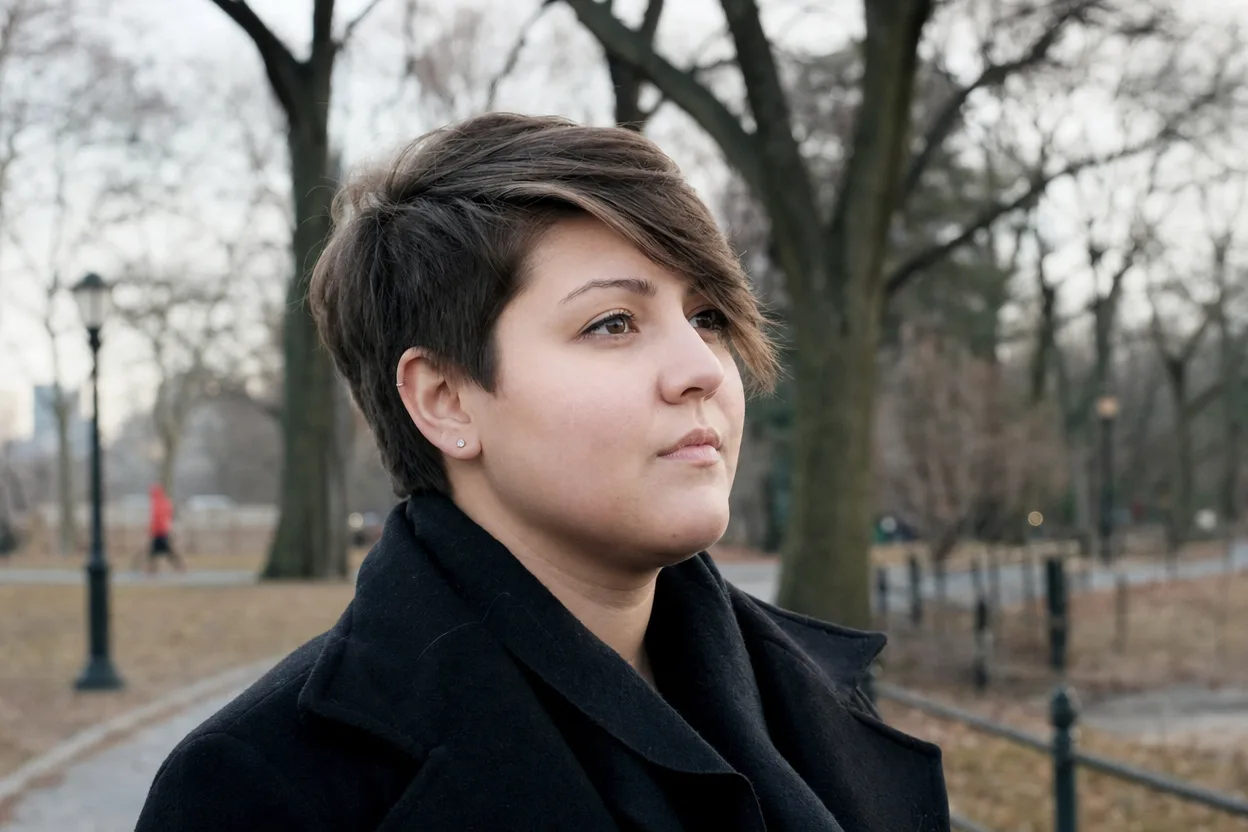Woman with a round face and mid brown hair in a park, her long side-swept fringe falling diagonally past her eyebrow from a deep side part
