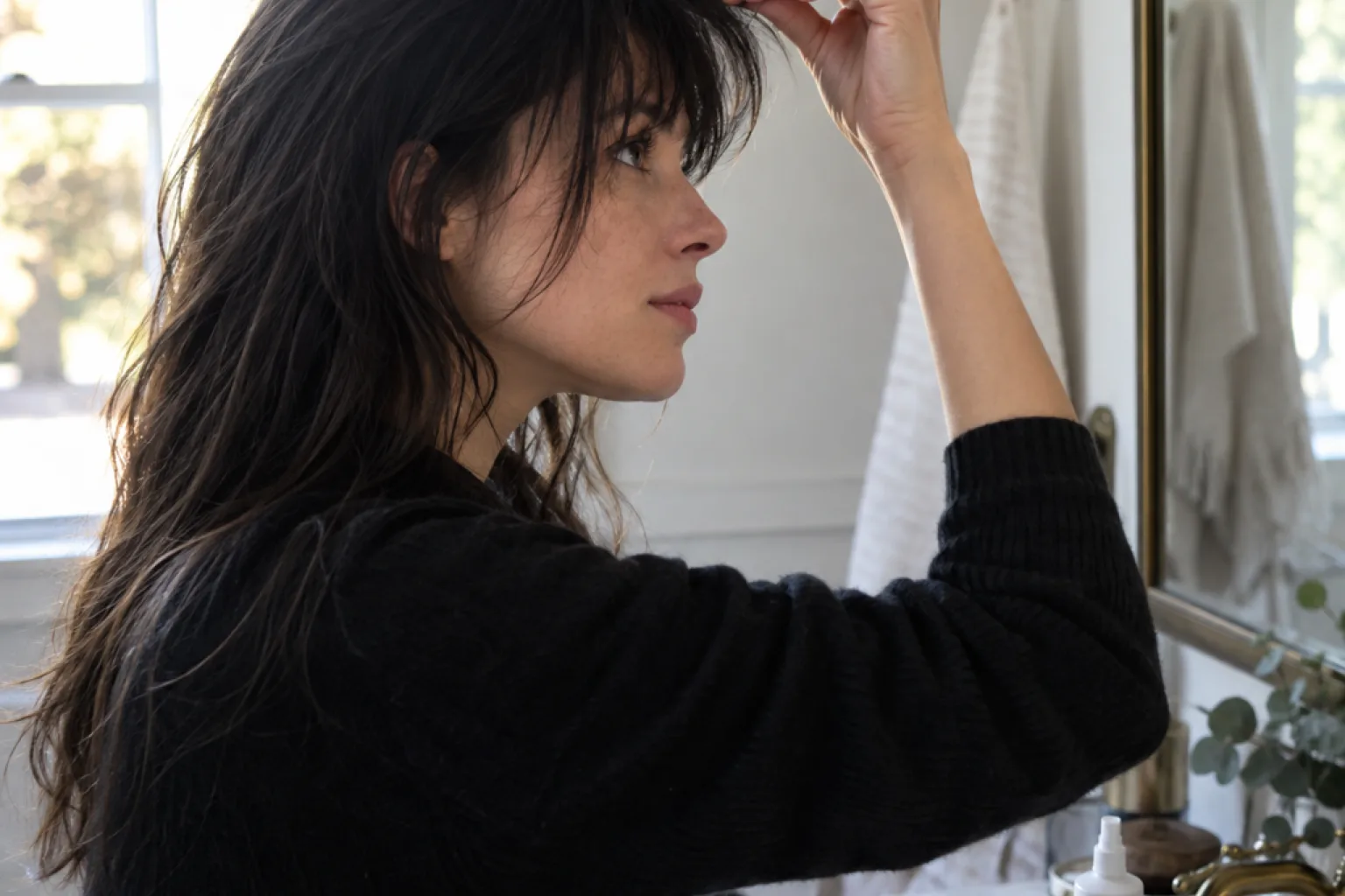 Woman with black hair at a bathroom mirror pinching her bangs between her fingers, a texture spray bottle on the basin beside her