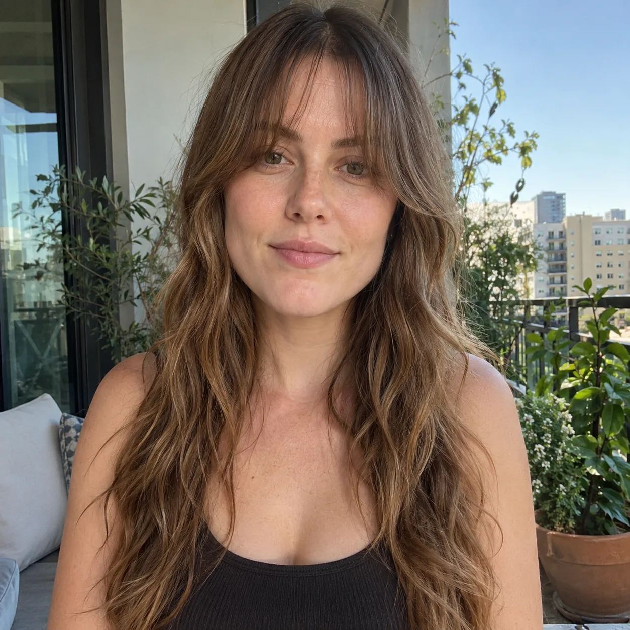 Woman with long wavy light brown hair on a sunny balcony, soft see-through curtain bangs with feathered ends