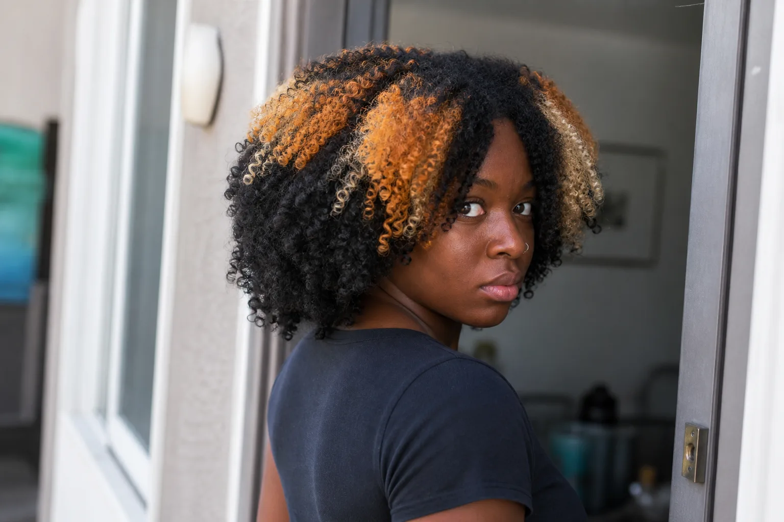 Woman with shoulder length coils in a doorway, near black base with copper and pale blonde clumps through the crown and front