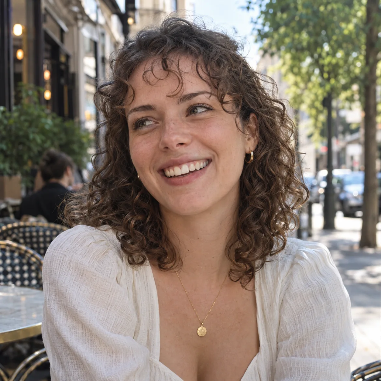 Woman with shoulder-length curly mid brown hair laughing at an outdoor cafe, a few loose curls falling over her forehead as a curly wispy fringe