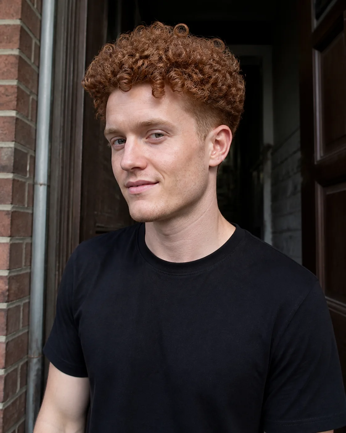Young auburn haired man in a brick doorway with defined springy curls standing tall on top while the sides are clipped close so the head is narrow at ear level