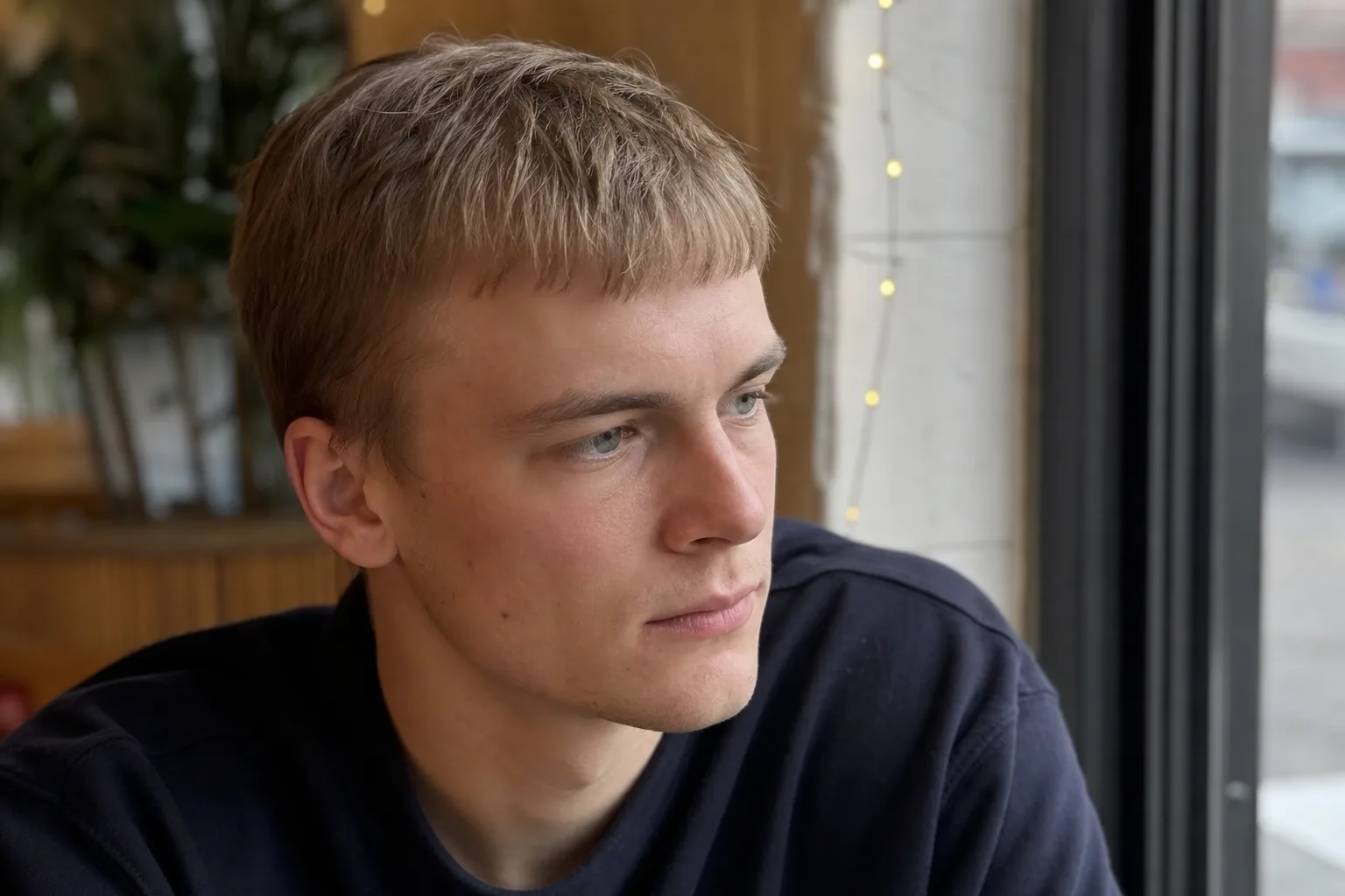 Young man by a cafe window with fine dirty blonde hair cut short and combed forward into a jagged fringe, the hair lying flat and close to the head
