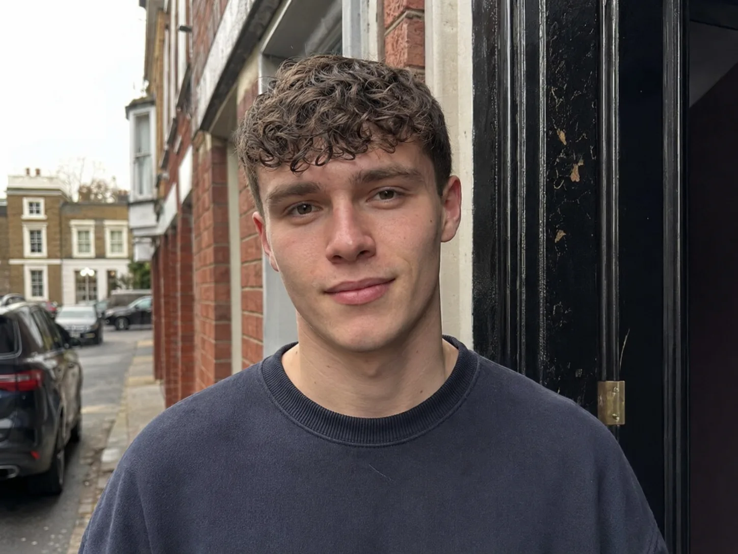 Young man in a brick doorway with a short curly crop and a curly fringe falling on his forehead
