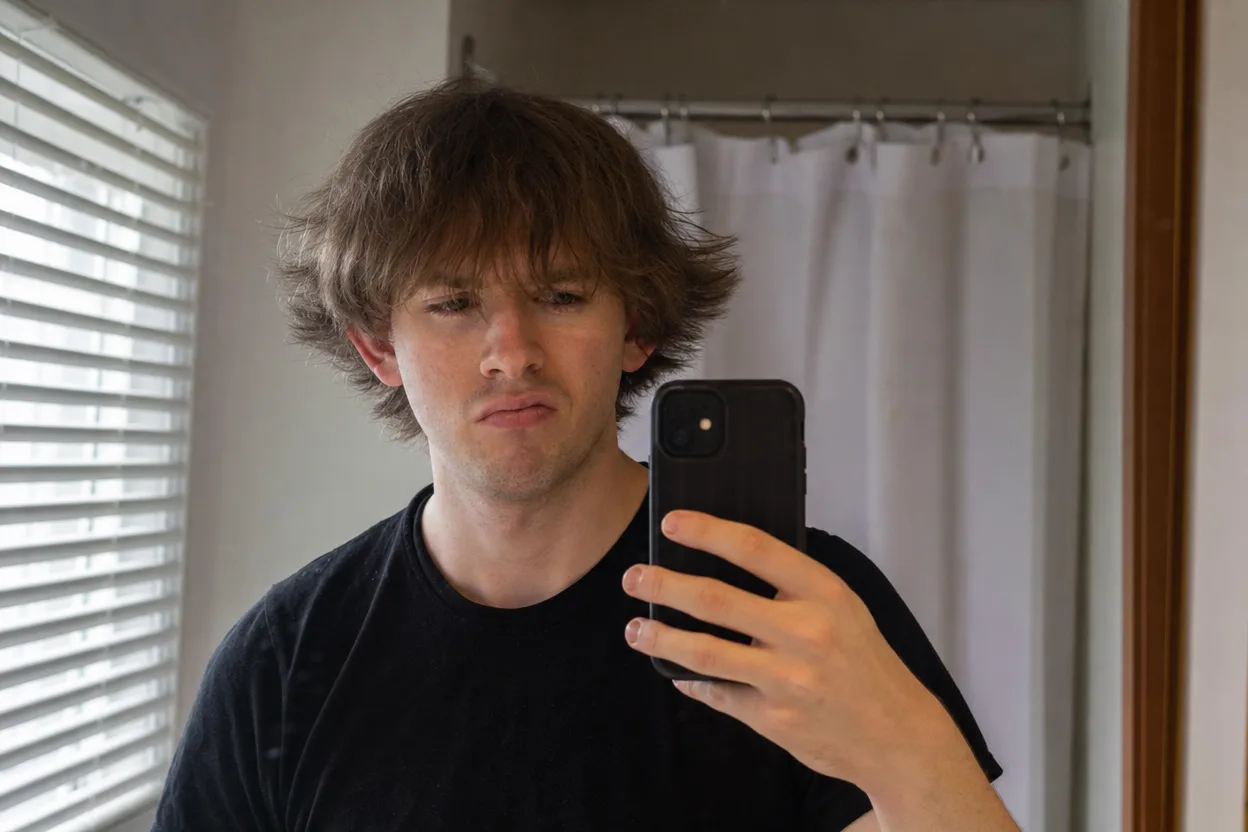 Young man taking a mirror selfie with unshaped three-inch hair puffing out over his ears and hanging into his eyes