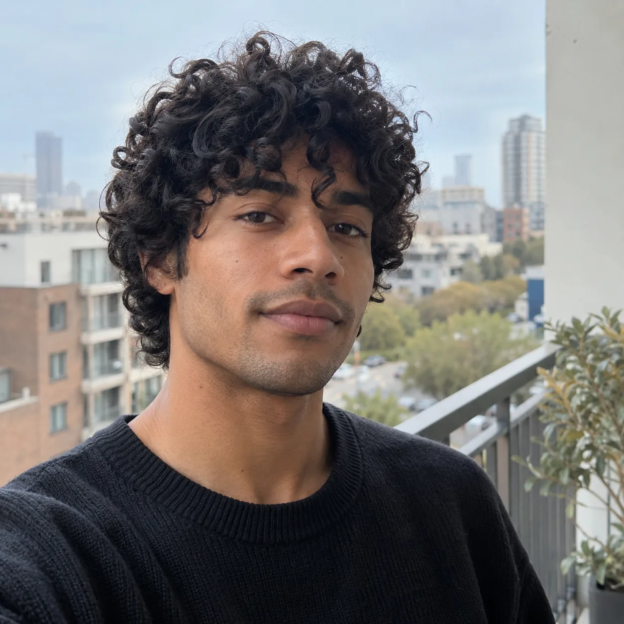 Young man with a curly black shag haircut and curls falling onto his forehead, on an apartment balcony